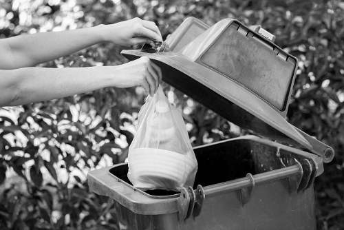 A person disposing of household waste into a black plastic bin with an open lid, holding a plastic shopping bag filled with white ceramic dishes. The bin is situated outdoors on a paved area, with a backdrop of foliage and bushes. The person's arms are visible and they are wearing short sleeves; the bin is positioned on the ground, with the lid propped open to facilitate waste disposal. The scene depicts routine rubbish collection which could be part of a commercial waste removal service, such as those offered by Commercial Waste Wembley, serving the Wembley area in northwest London. The image emphasizes the act of waste disposal, possibly within a domestic or small commercial setting, with a focus on maintaining cleanliness and proper waste segregation. Bright natural lighting highlights the textures of the plastic bin, the smooth surface of the ceramic dishes, and the surrounding greenery, illustrating a typical scene of rubbish disposal relevant to waste management services in the Wembley postcode region.
