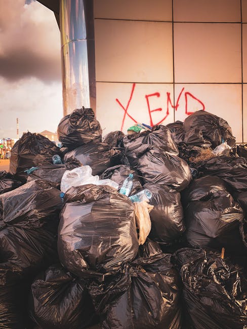 A large pile of black garbage bags filled with waste is stacked against an exterior wall of a building, with some bags appearing to be overfilled and bulging. Several plastic water bottles, some parti