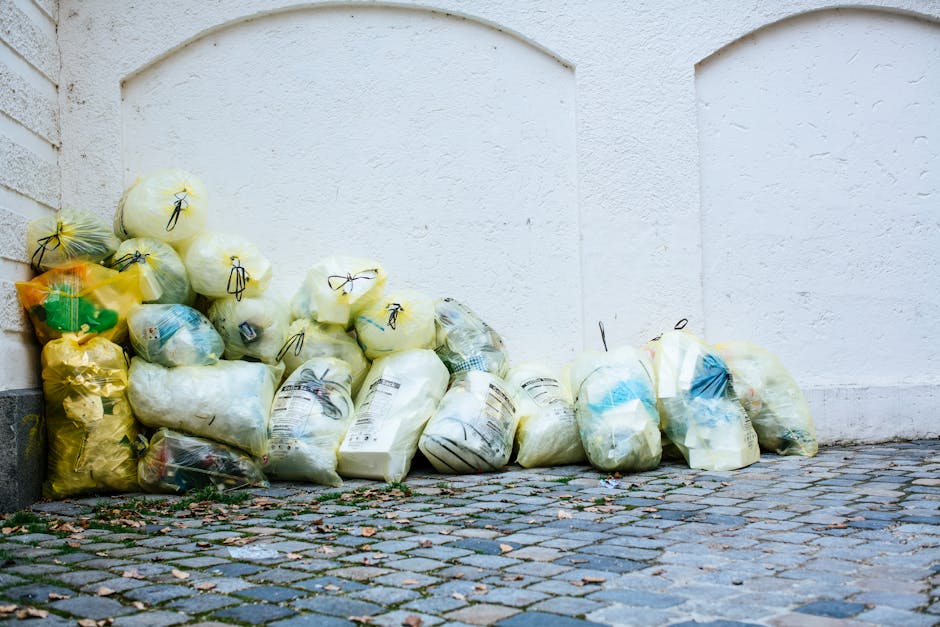 A large pile of black garbage bags filled with waste is stacked against an exterior wall of a building, with some bags appearing to be overfilled and bulging. Several plastic water bottles, some parti