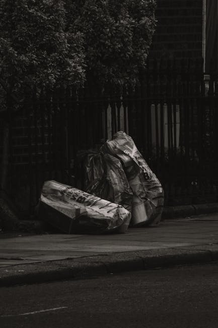 A collection of overflowing rubbish bins and scattered waste on a paved urban sidewalk, including large black and transparent plastic garbage bags, flattened cardboard boxes, discarded paper and packa